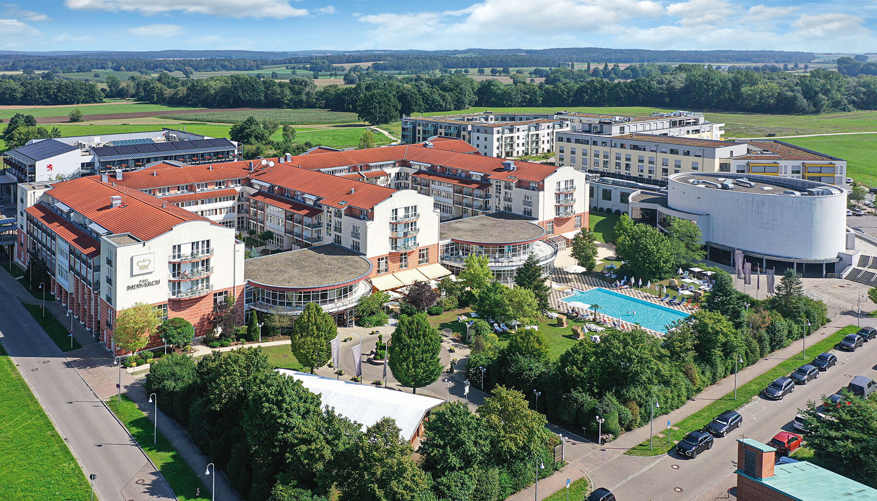 Das Hotel von oben: mehrere Flügel mit roten Ziegeldächern, ein runder Kuppelbau und ein Freibad im Grünen, ringsum Felder